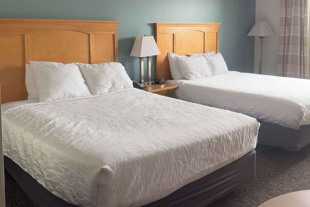 Quarto de hotel com duas camas, cabeceiras de madeira, roupa de cama branca, luminária de cabeceira e mesa de cabeceira.