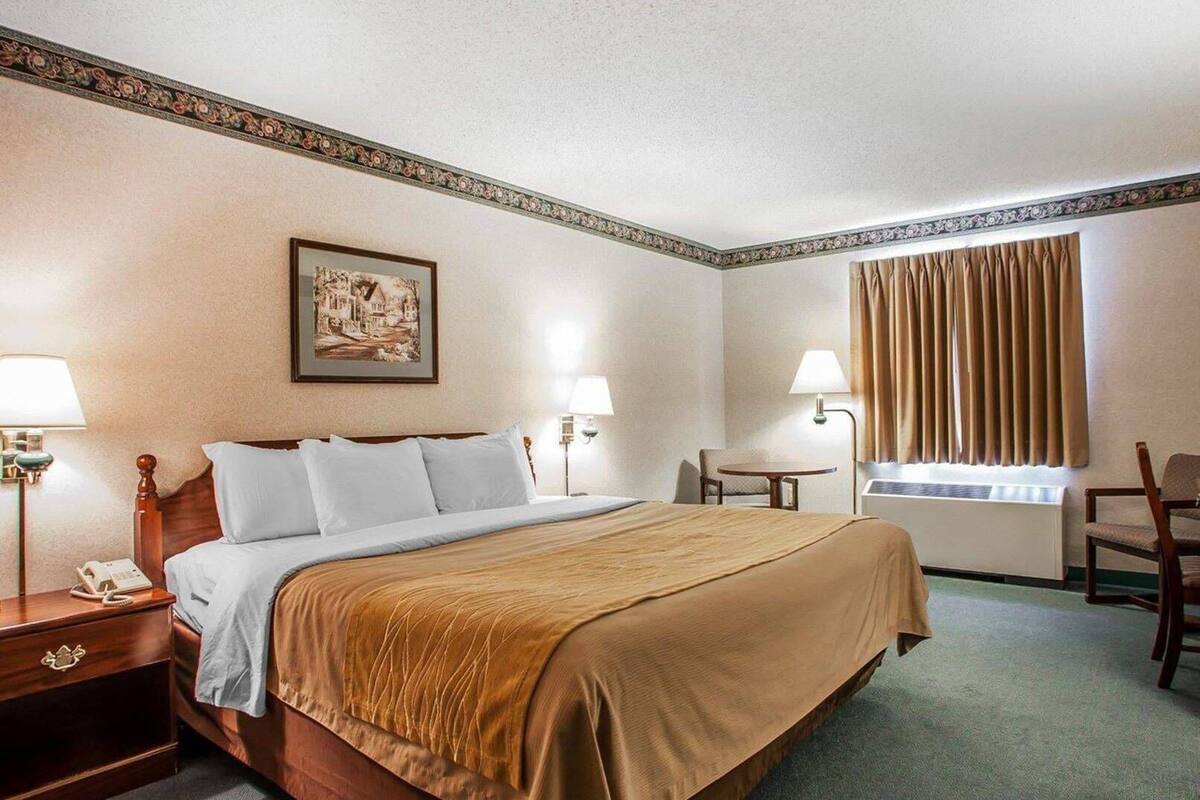 Habitación de hotel con cama, lámparas de noche, una silla, una mesita, un cuadro y ventana con cortinas.