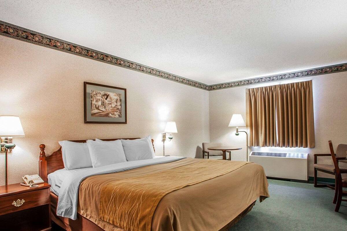 Habitación de hotel con una cama grande, dos mesitas de noche, un escritorio, una silla, una ventana con cortinas y un cuadro enmarcado en la pared.
