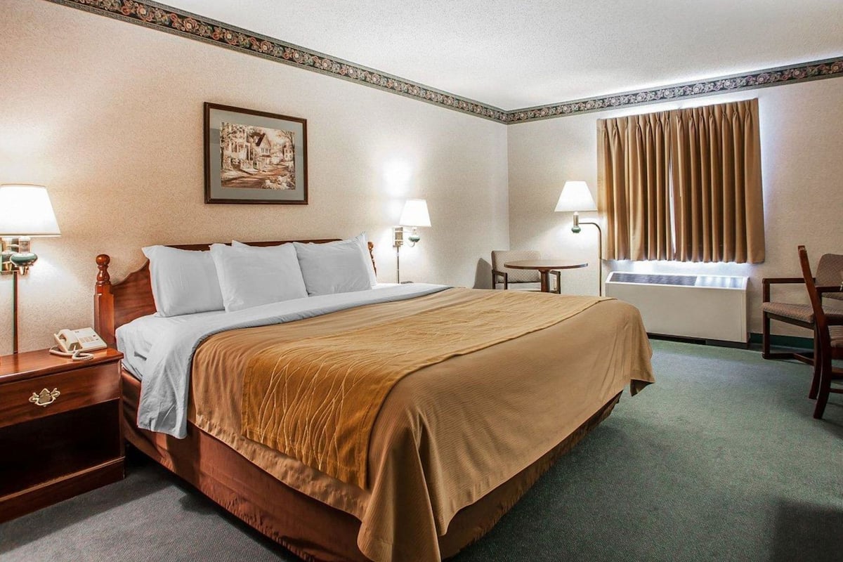 Una habitación de hotel con cama, mesitas de noche, escritorio, silla, ventana con cortinas e imagen colgada en la pared.