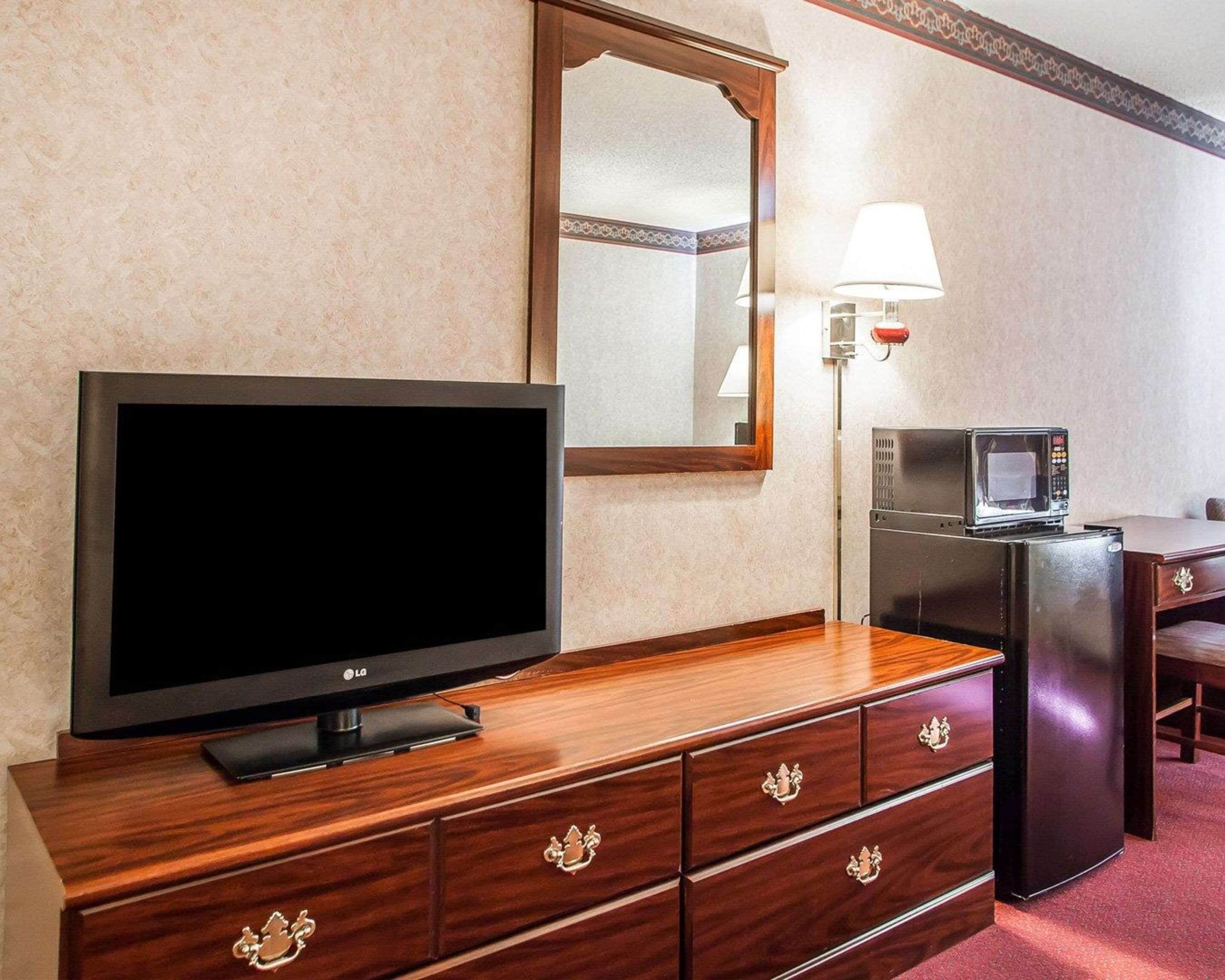 A hotel room with a wooden TV stand, a flat-screen TV, a refrigerator, a microwave, a lamp, and a mirror.