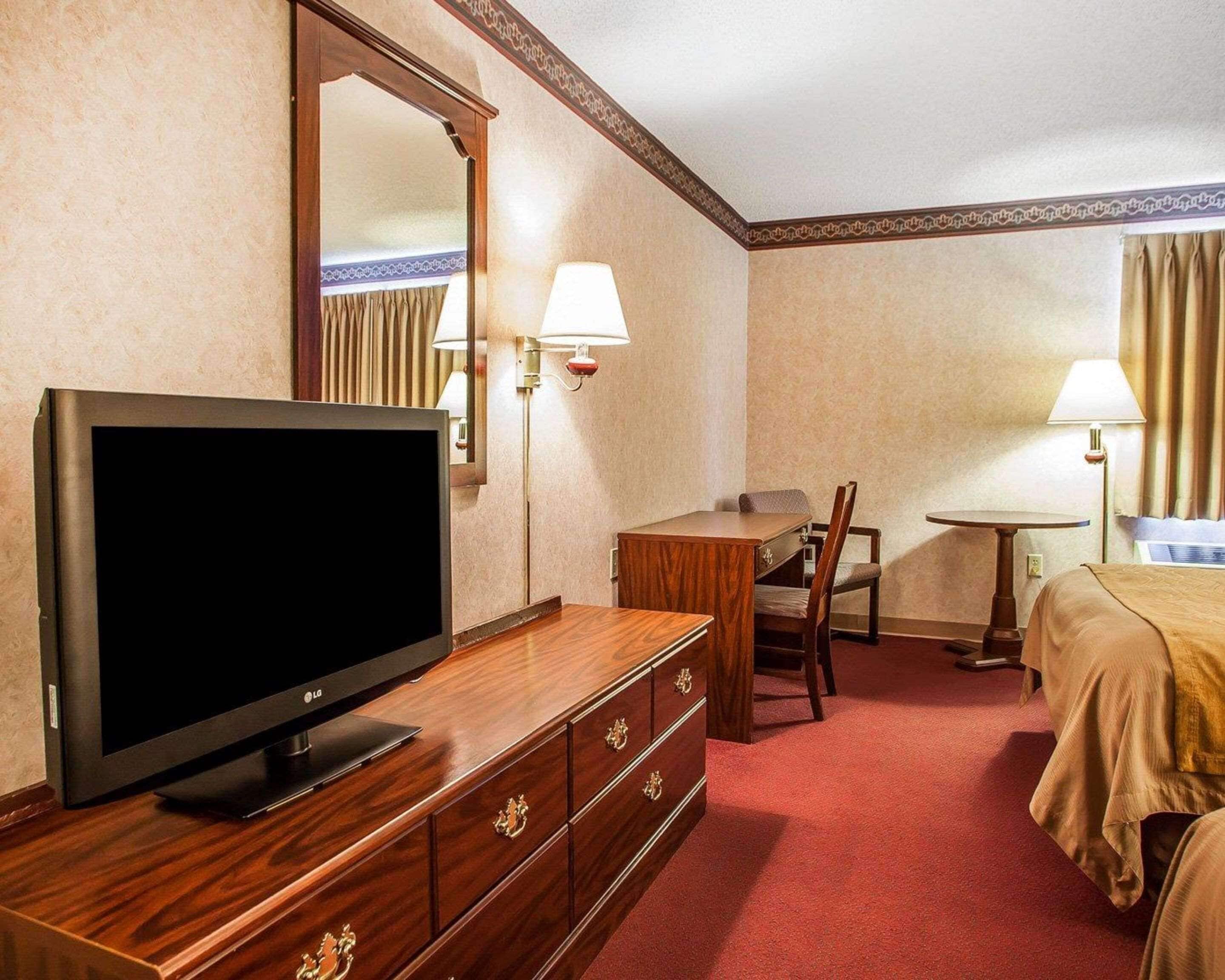 A hotel room with a TV on a wooden dresser, a desk with a chair, a bed, and two bedside lamps.
