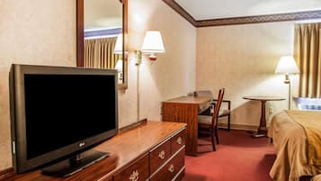 A hotel room with a TV on a wooden dresser, a desk with a chair, a bed, and two bedside lamps.