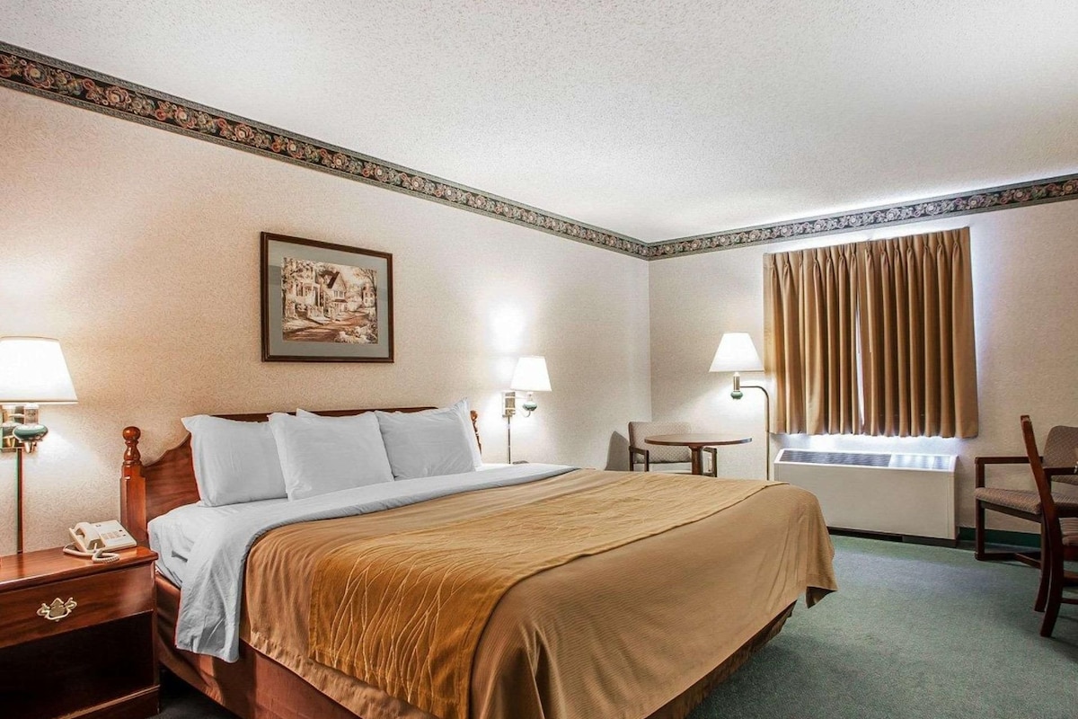 A hotel room with a bed, bedside tables, a desk, a chair, a window with curtains, and a wall-mounted lamp.