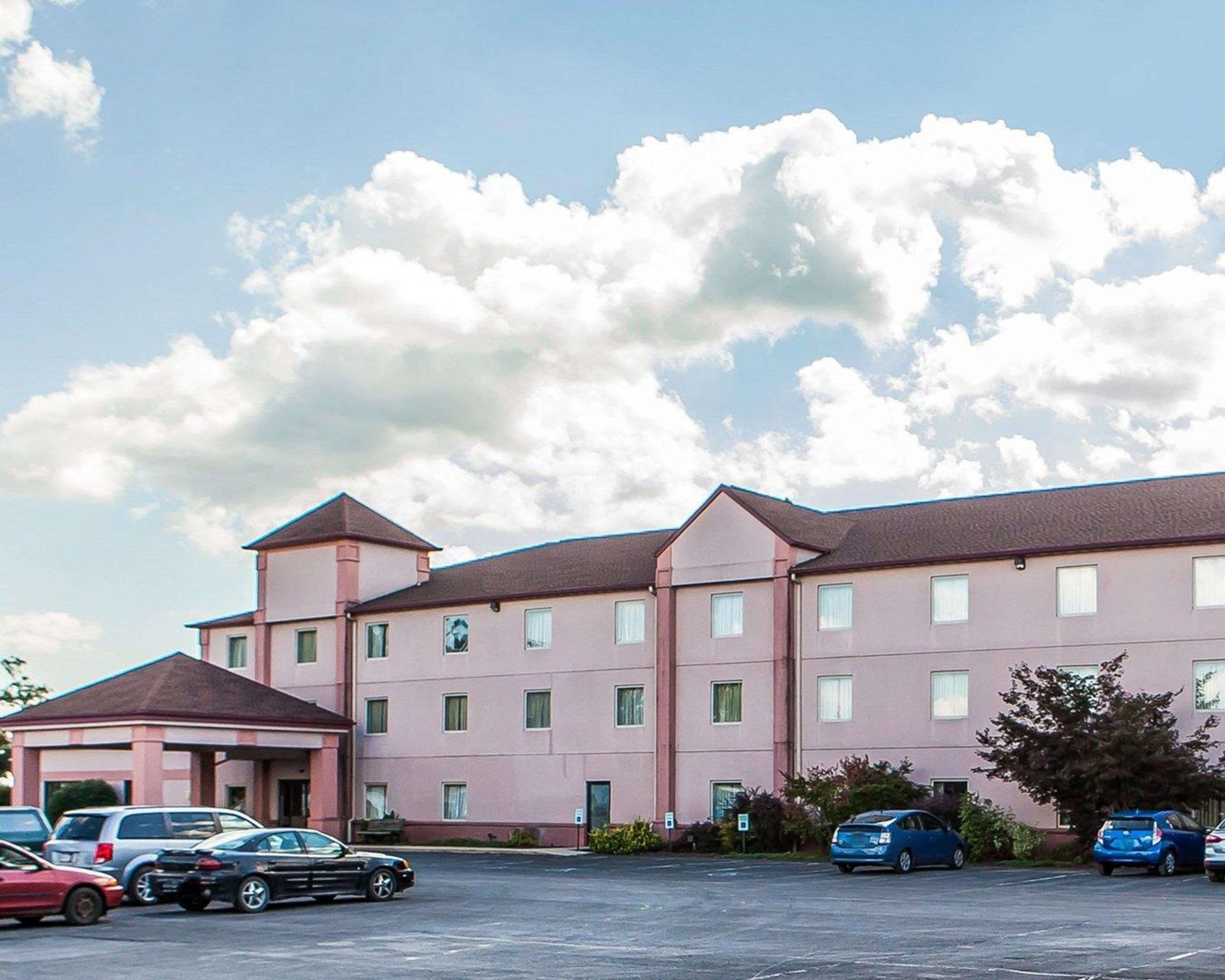 Un hotel de varios pisos con entrada principal, estacionamiento y cielo despejado con nubes.