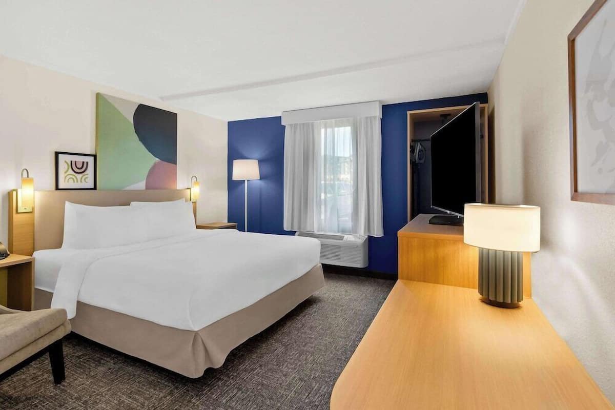 A hotel room with a bed, bedside tables, a lamp, a window with curtains, and a wall art piece.