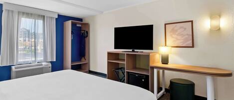 A hotel room with a bed, a television, a desk with a lamp, a trash bin, and a wardrobe.