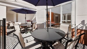 Un patio con una mesa redonda y sillas, un gran parasol y un ventanal de vidrio que da a un espacio interior.