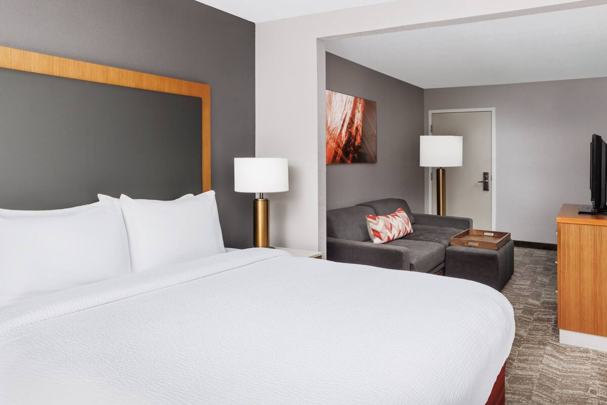 A hotel room with a large bed, a grey sofa, a wooden desk, and a television.