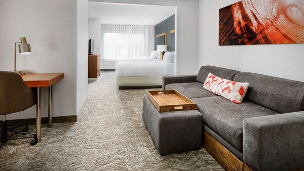 Una habitación de hotel moderna con un sofá gris, una mesita de madera, un escritorio con lámpara y una cama grande con ropa de cama blanca.