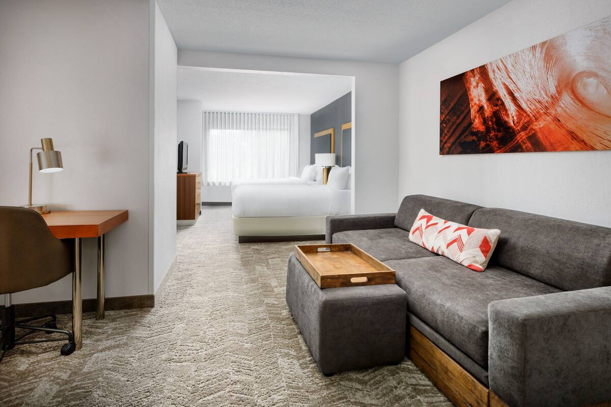 A modern hotel room with a grey sofa, a wooden side table, a desk with a lamp, and a large bed with white bedding.