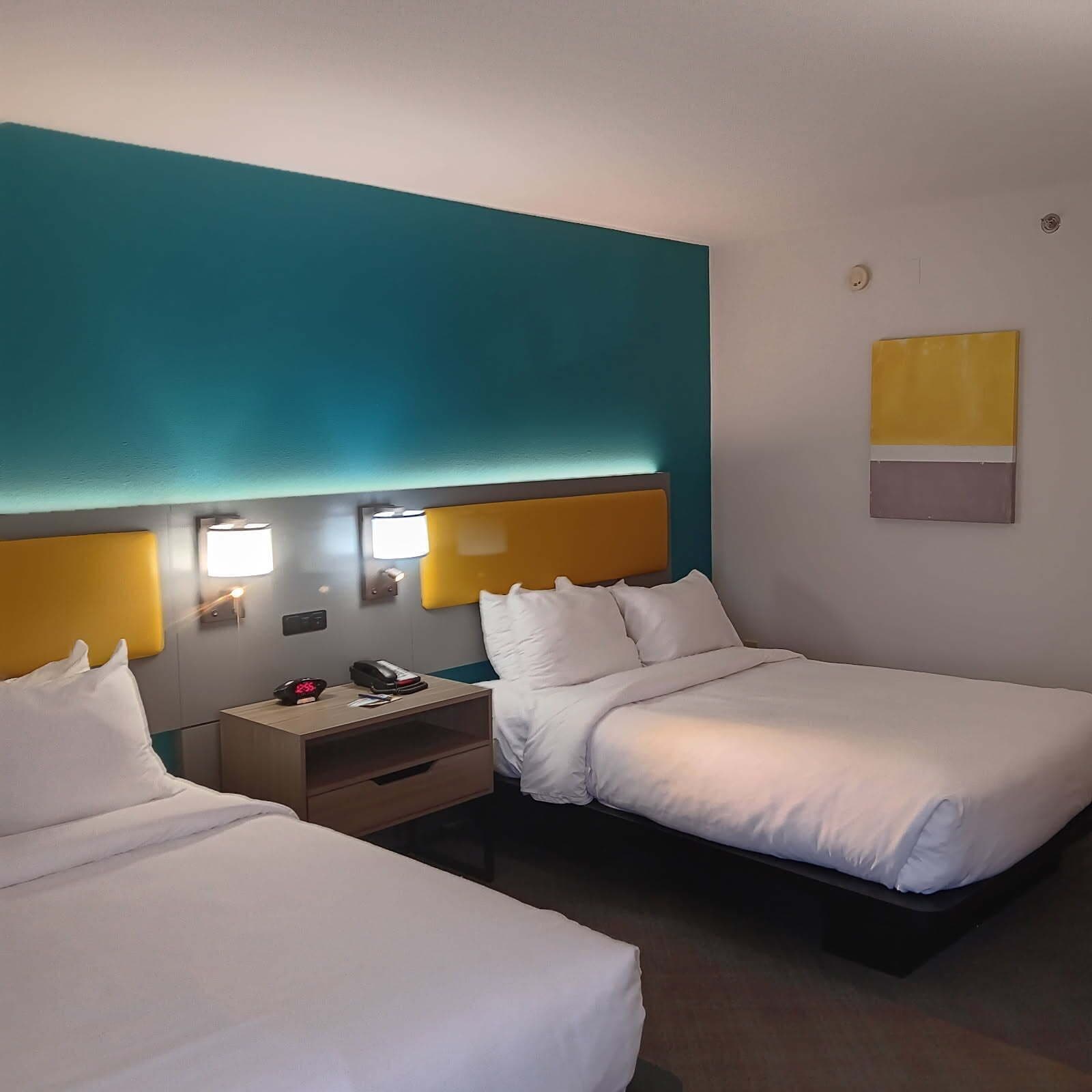 Una habitación de hotel con dos camas, una mesita de noche, una lámpara y una obra de arte colgada en la pared.