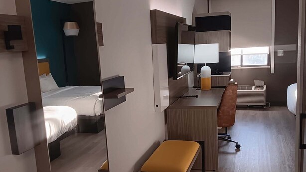 Une chambre d’hôtel avec un lit, un bureau, une chaise, un petit banc, un miroir et une fenêtre.