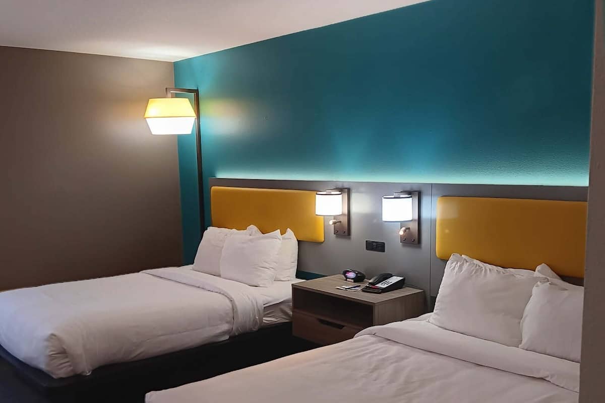 Ein Hotelzimmer mit zwei Betten, jedes mit weißer Bettwäsche und einem gelben Kopfteil. Nachttische mit Lampen und einer digitalen Uhr sind ebenfalls vorhanden.