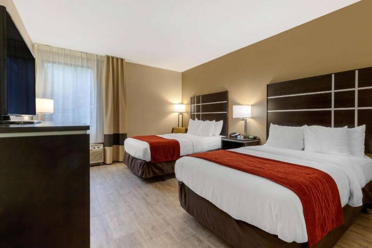 Ein Hotelzimmer mit zwei Betten, einem Schreibtisch, einer Lampe und einem großen Fenster mit Vorhängen.