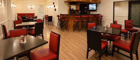 Comedor de hotel con sillas tapizadas rojas, mesas negras y una zona de bar con una pantalla de televisión.