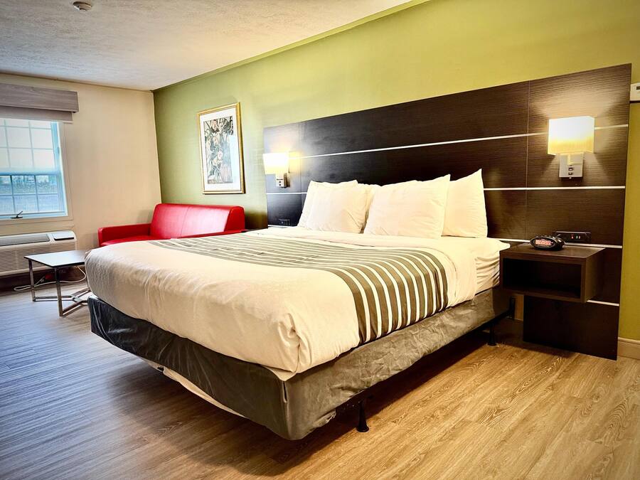 A hotel room with a large bed, a red chair, a small table, and a window with blinds.