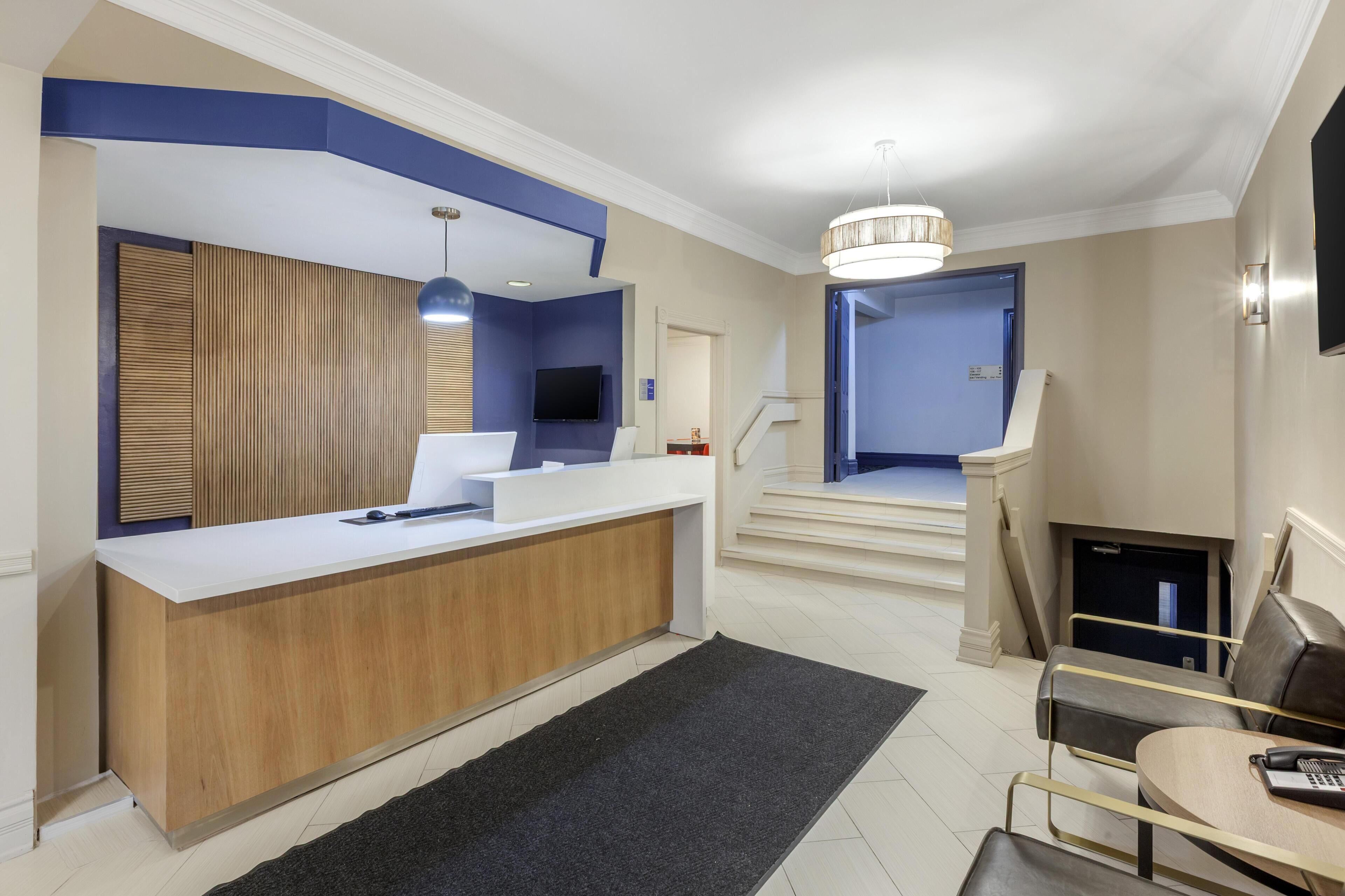 A hotel reception area with a wooden reception desk, a flat-screen TV, a chandelier, and a staircase leading to another floor.
