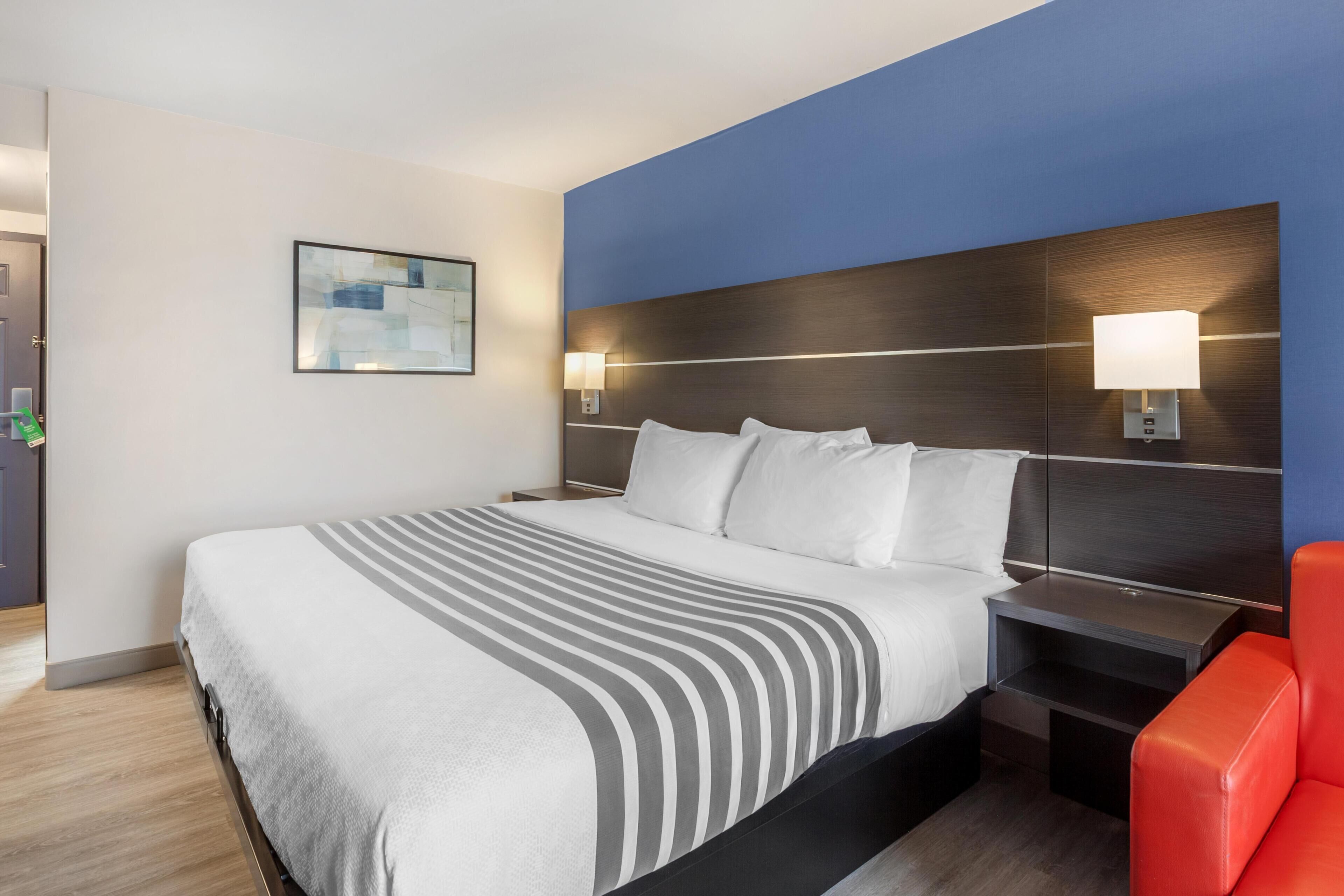 A hotel room with a large bed, bedside tables, a red chair, and a painting on the wall.