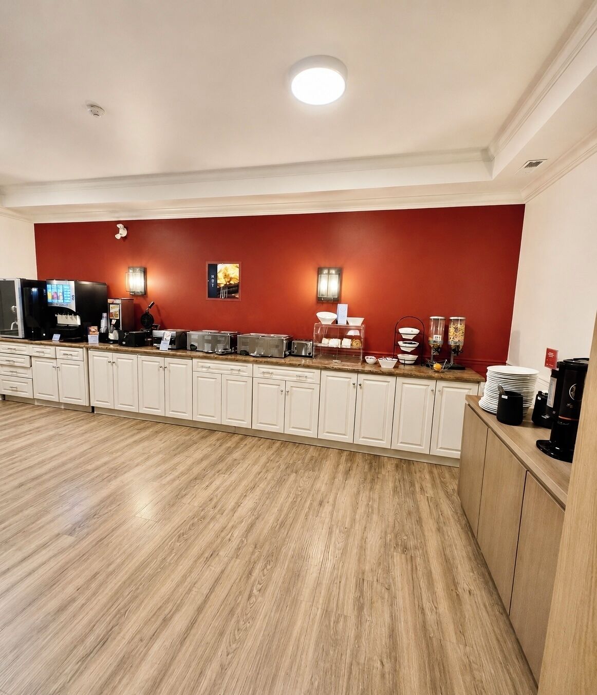 Una cocina de hotel con pared roja de acento, armarios blancos y suelo de madera.