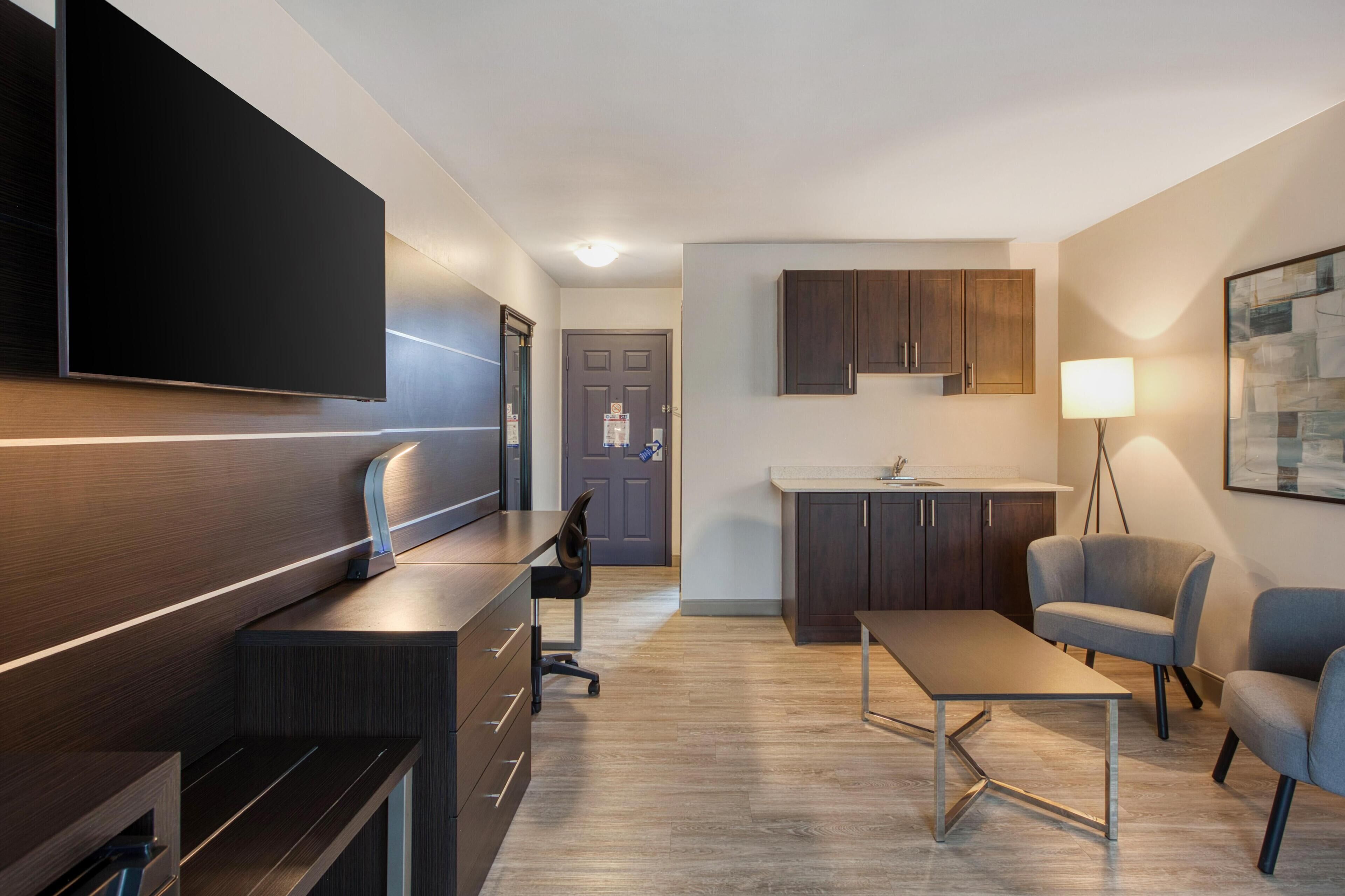 A modern living room with a flat-screen TV, a desk, a chair, a sofa, a lamp, and a kitchen area with cabinets.
