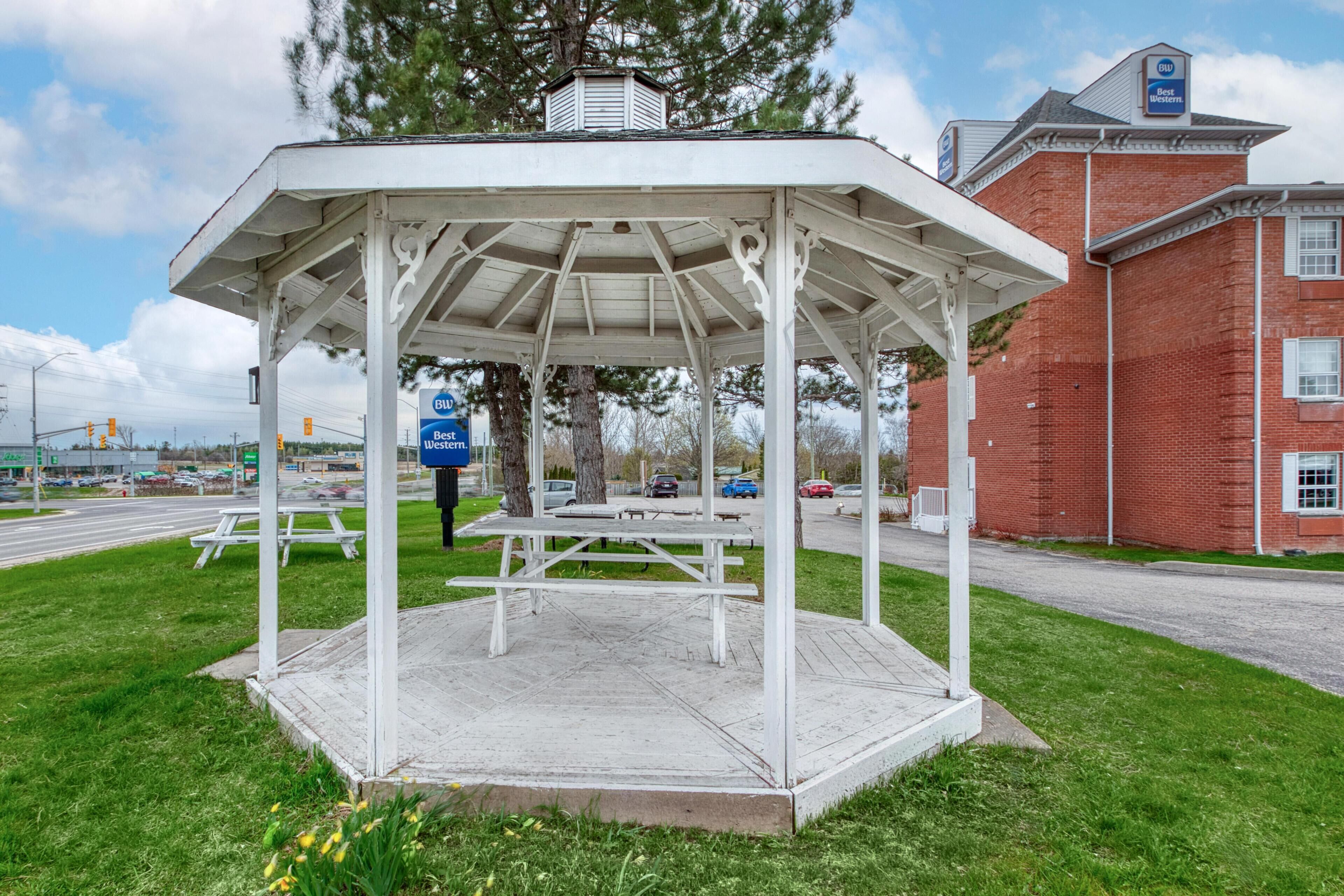A gazebo with a roof, surrounded by grass, and a brick building in the background.