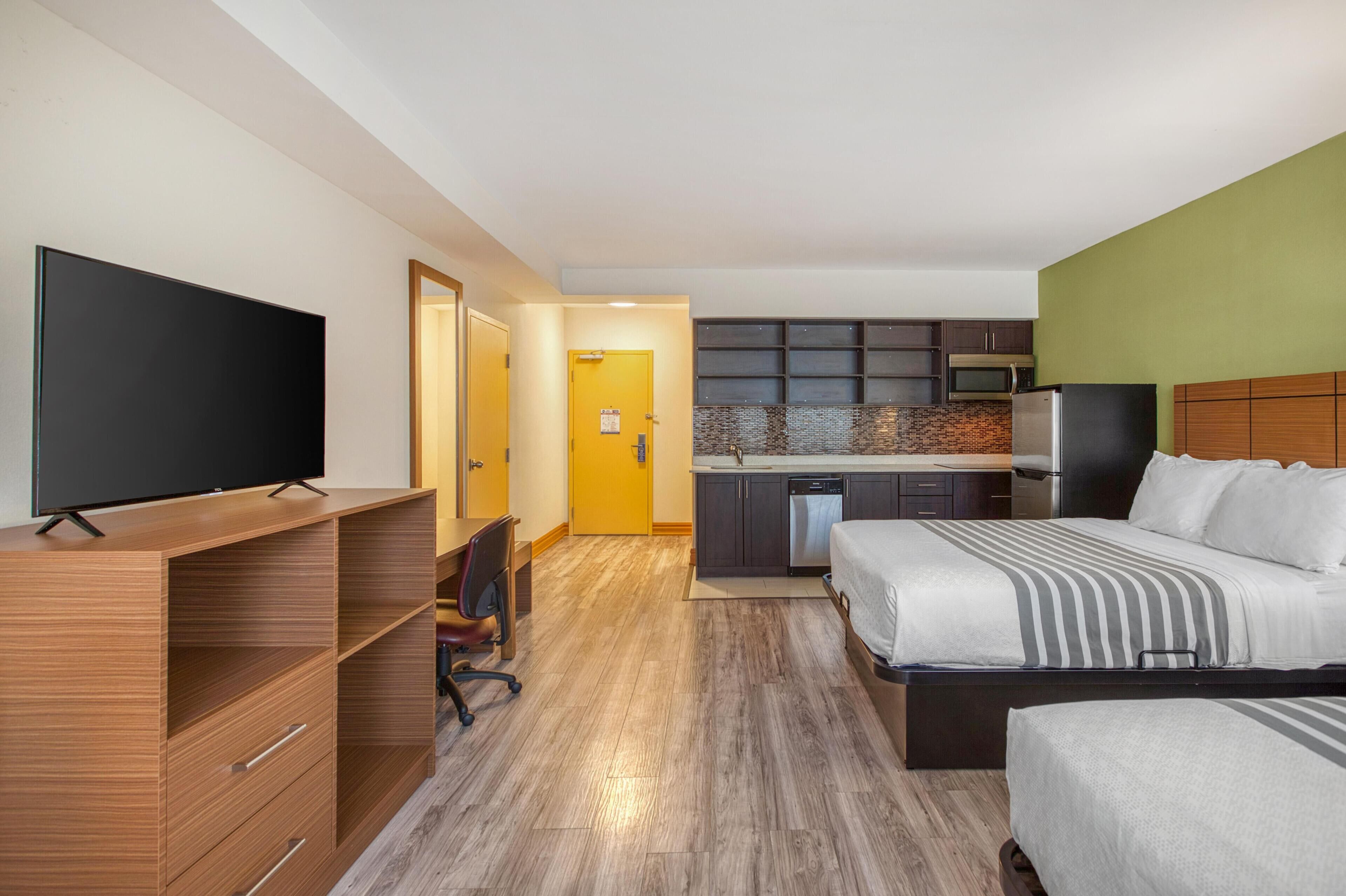 A hotel room with a TV, a bed, a desk, and a kitchenette.