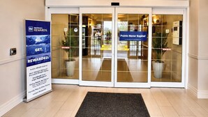 L’entrée d’un hôtel au design moderne, avec une grande porte vitrée, un paillasson noir et un panneau indiquant les avantages offerts.