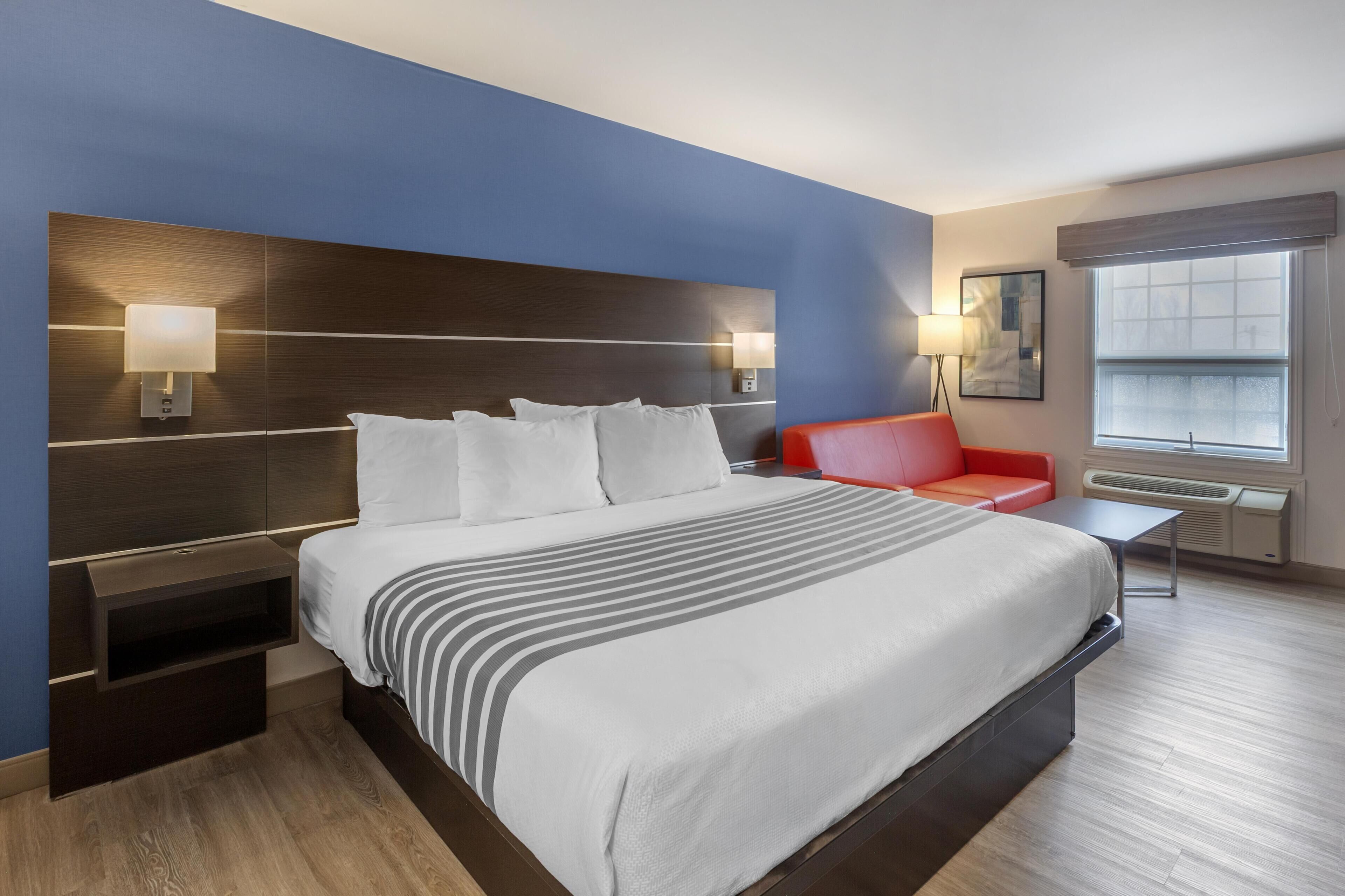 Habitación de hotel con una cama grande, un sofá rojo, una mesita pequeña y una ventana con persianas.