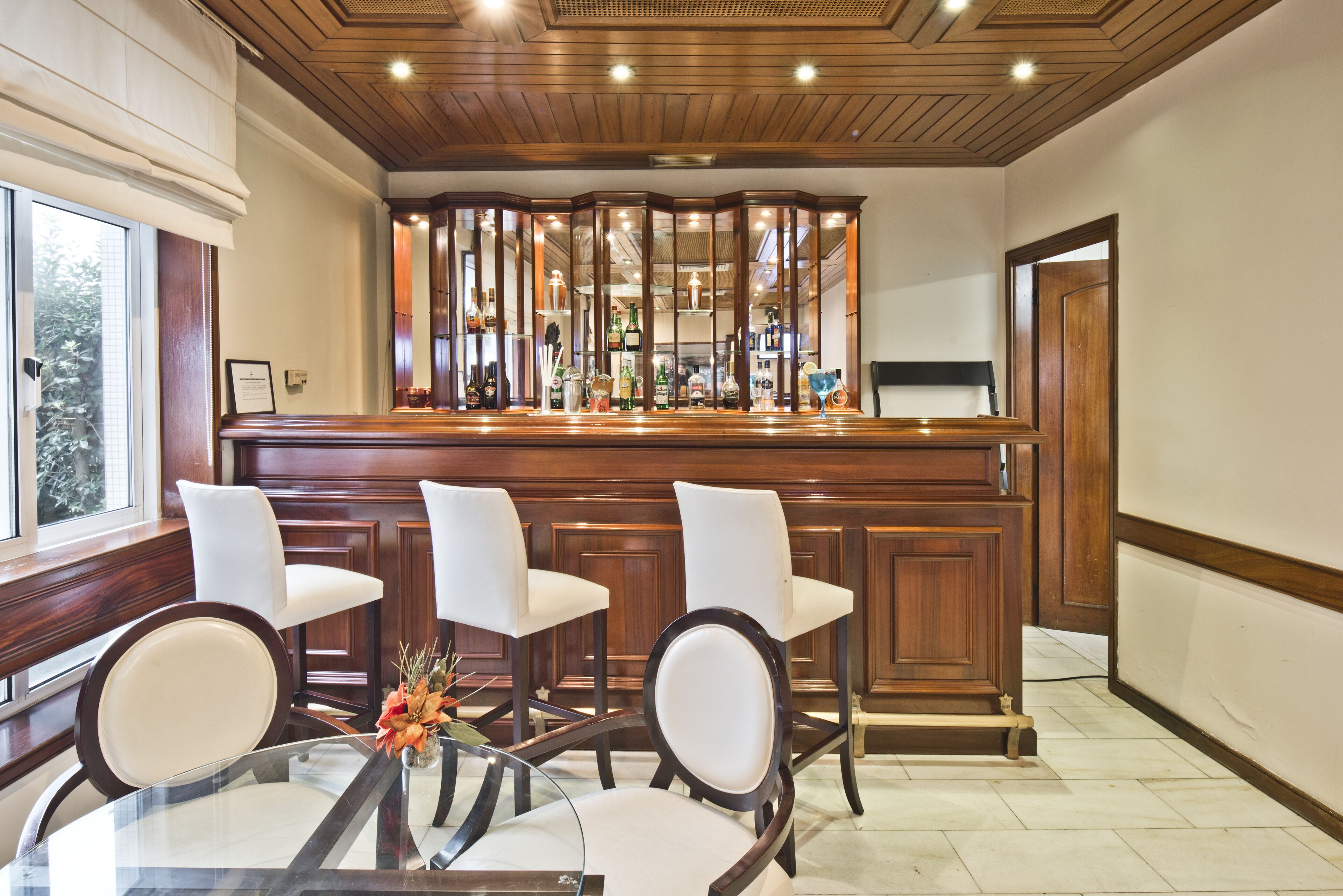 Zona de bar con mostrador de madera, sillas blancas tapizadas y una mesa de vidrio con un florero.