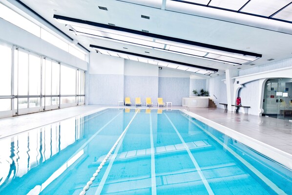 Uma grande piscina interior com água azul cristalina, cadeiras de descanso amarelas e um design moderno no teto.