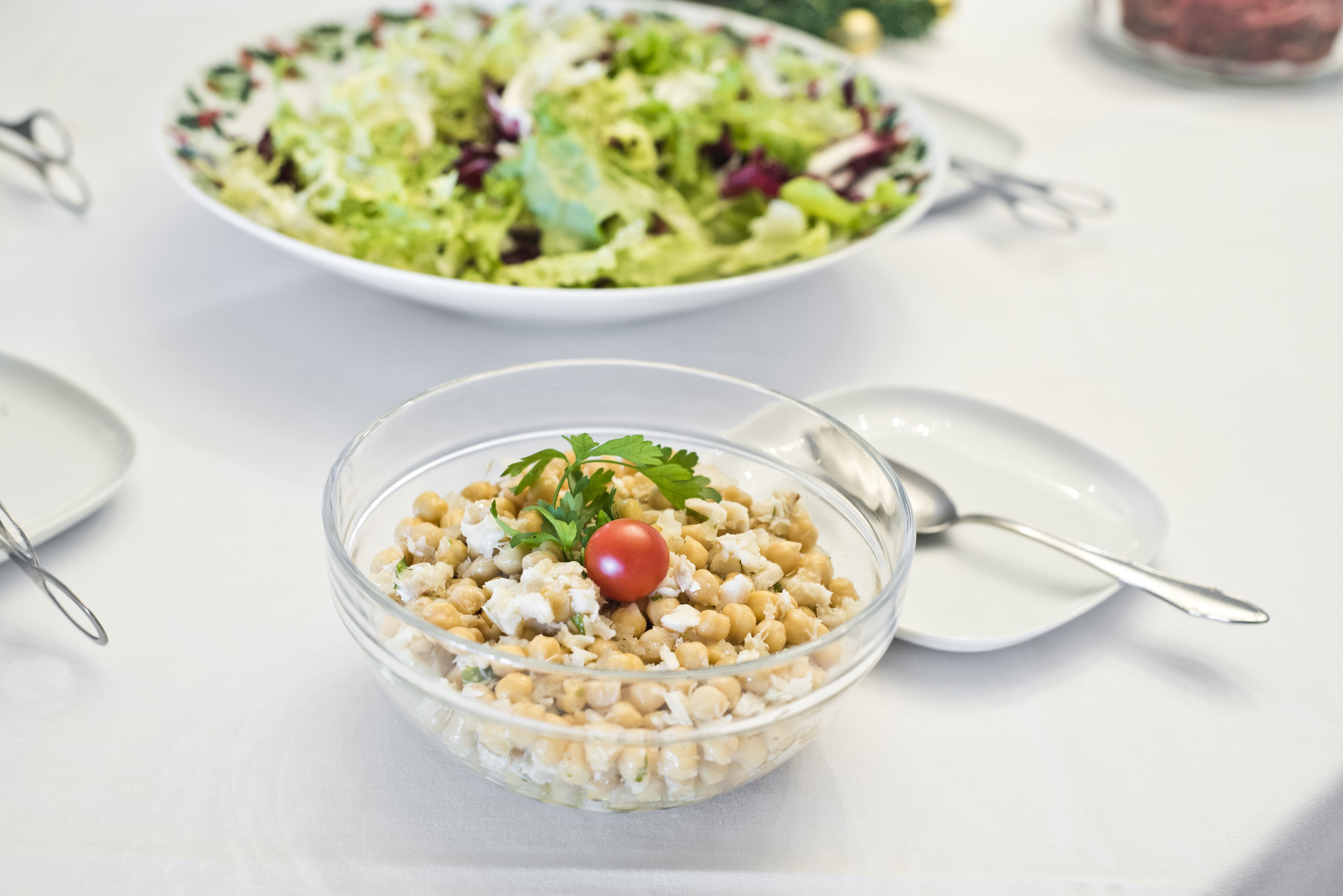 Un tazón de vidrio transparente con ensalada de garbanzos, adornado con un tomate cherry y perejil, sobre una mesa blanca junto a una cuchara y un plato.