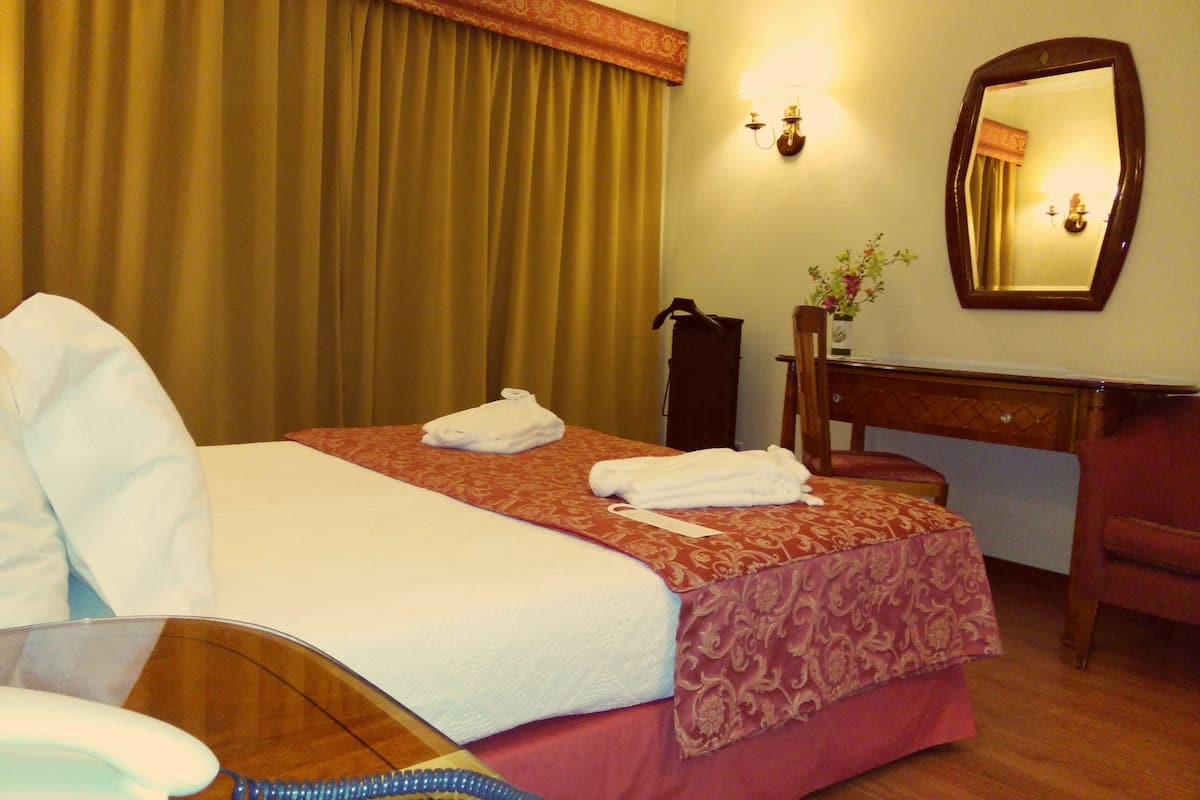 Une chambre d’hôtel avec un lit, un bureau, une chaise, un miroir et un vase avec des fleurs.