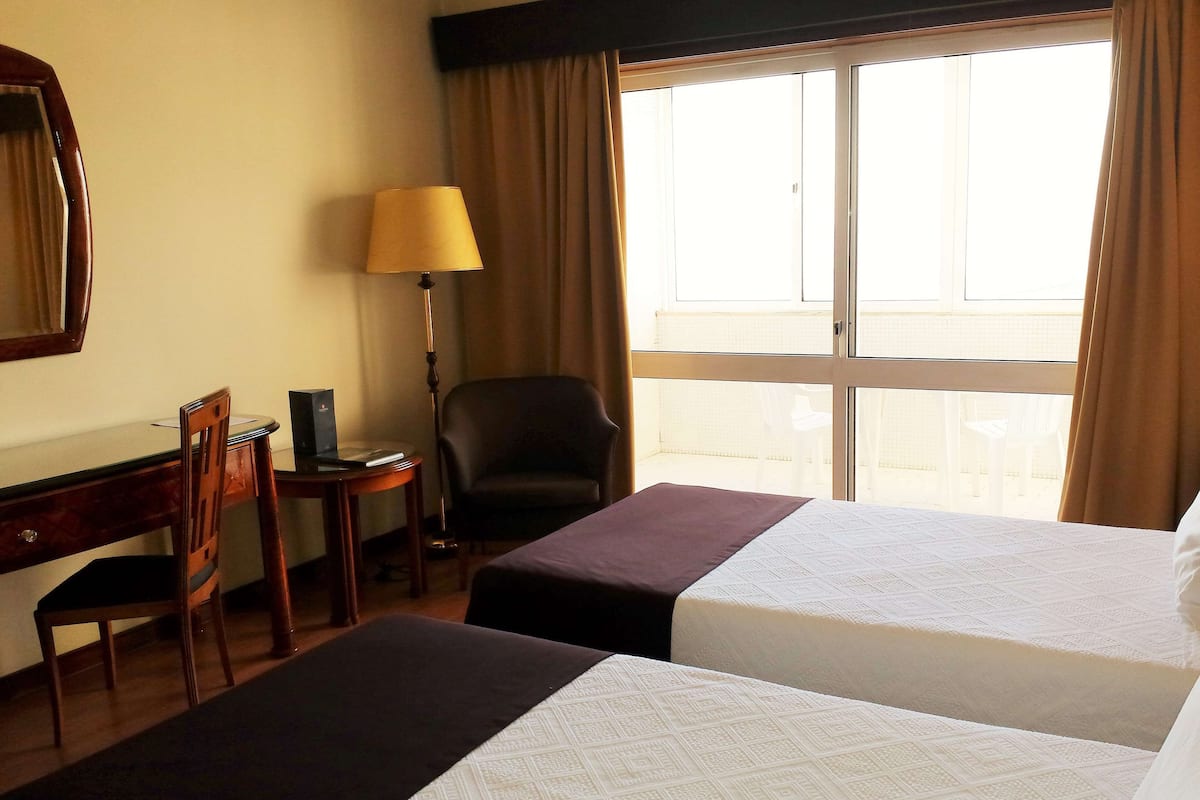 Une chambre d’hôtel avec deux lits, un bureau, une chaise, une lampe et une fenêtre avec des rideaux.