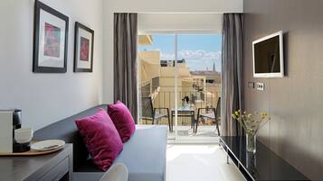 Une chambre d’hôtel moderne avec un canapé gris, une petite table sur laquelle se trouvent une tasse et une soucoupe, un téléviseur fixé au mur et un balcon offrant une vue sur les bâtiments et les montagnes.