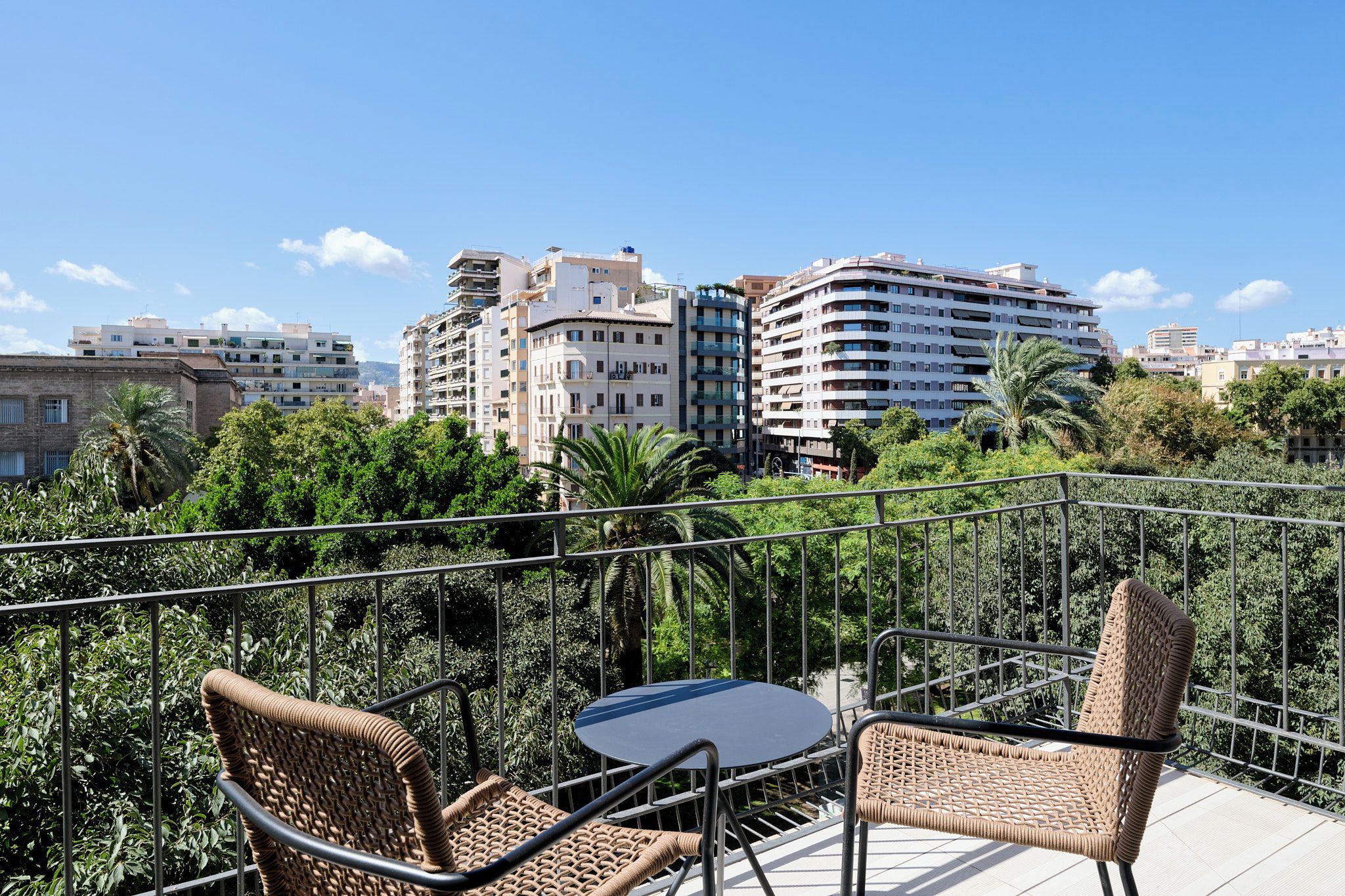 Un balcón con mobiliario de mimbre y una mesa, con vistas a un paisaje urbano con rascacielos y áreas verdes.