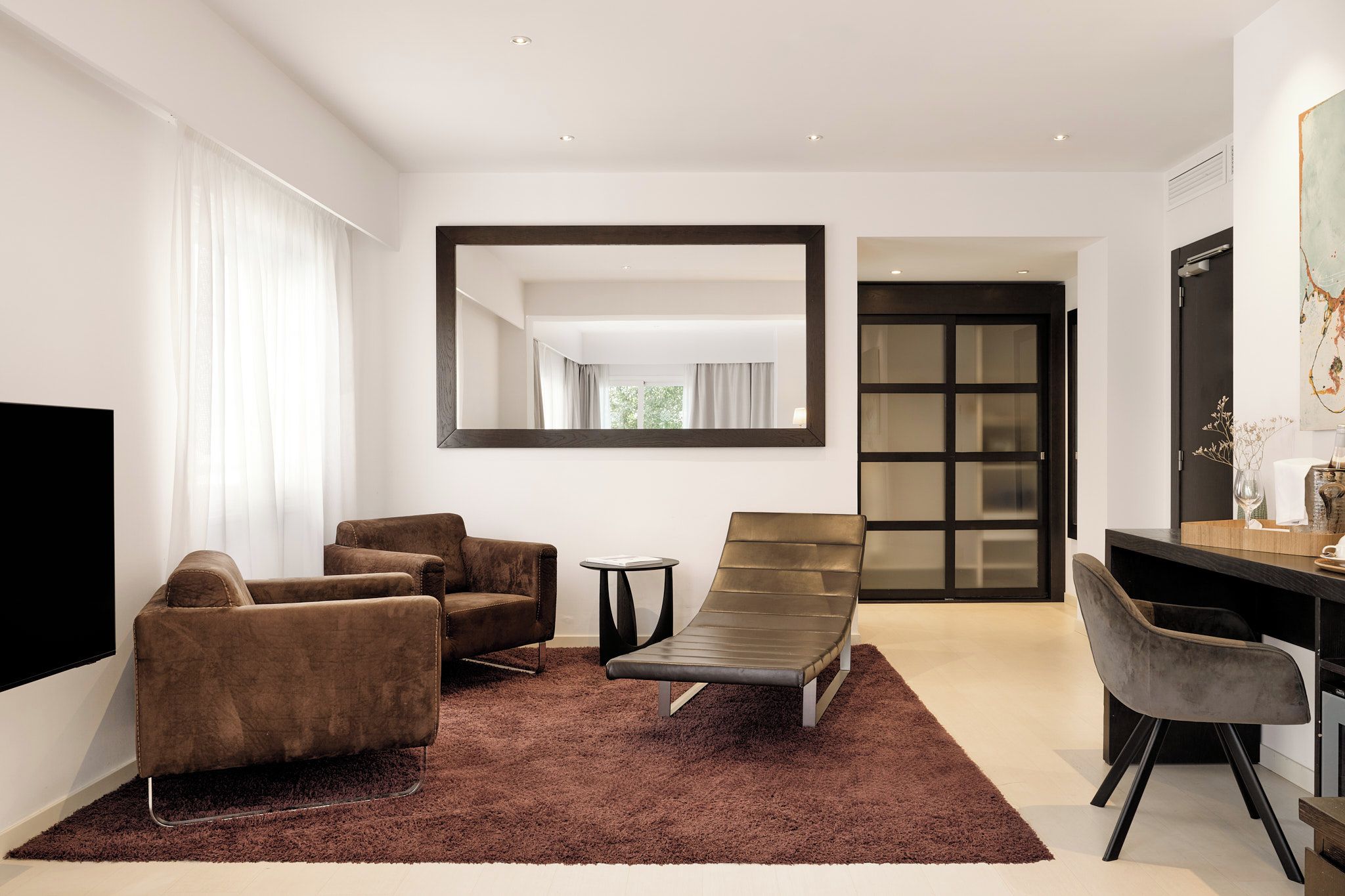 A modern living room with a large flat-screen TV, two brown sofas, a single chair, a small side table, and a large mirror on the wall.