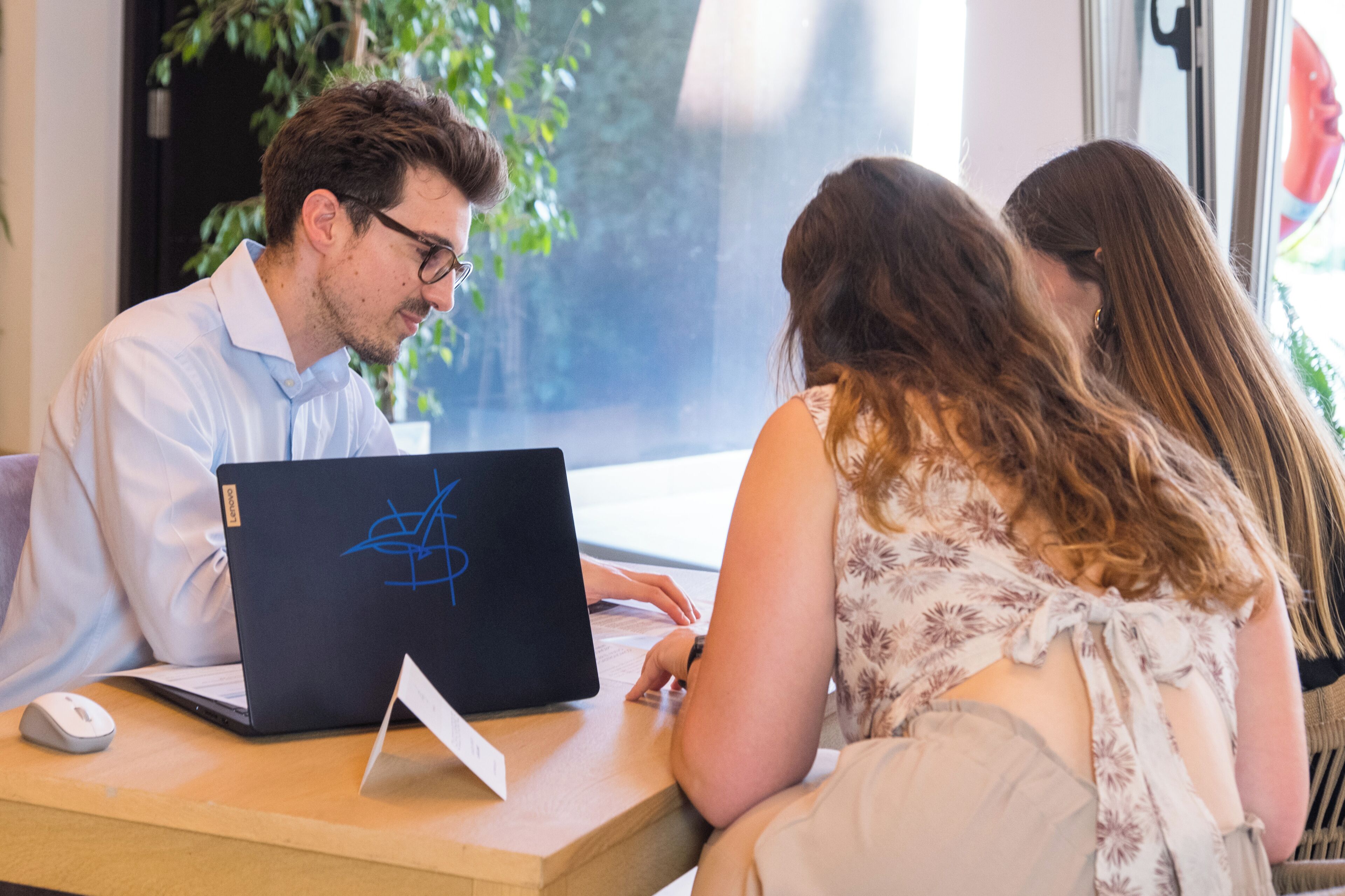 Une personne utilise un ordinateur portable sur une table en bois, avec une souris blanche, dans une pièce dotée d’une fenêtre et d’une plante en arrière-plan.