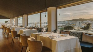 Ein Restaurant mit gedeckten Tischen, großen Fenstern mit Blick auf die Stadt und hängenden Pendelleuchten.