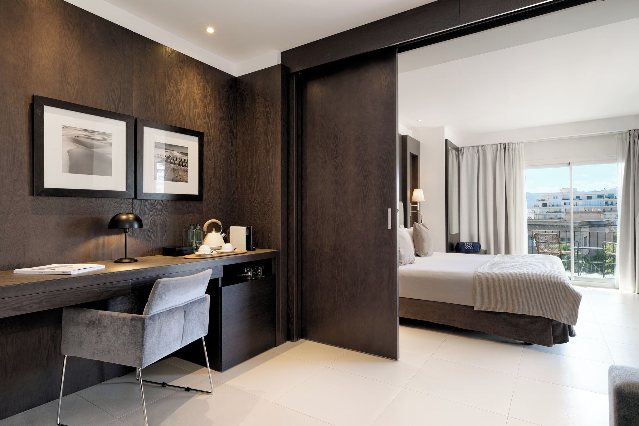 A modern hotel room with a dark wood panel wall, a desk with a chair, a bed with a view, and a large window.