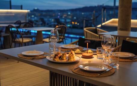 Una mesa de comedor lista con platos, vasos y cubiertos, con vistas a un paisaje urbano al anochecer.