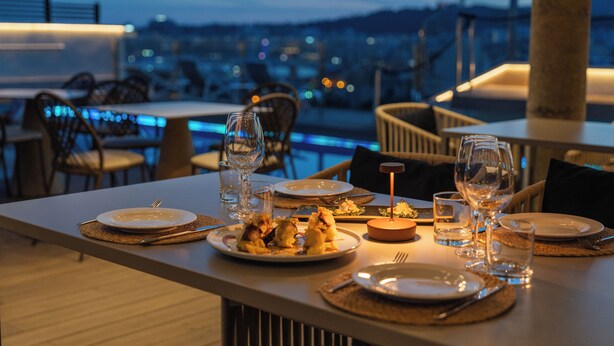 Una mesa de comedor lista con platos, vasos y cubiertos, con vistas a un paisaje urbano al anochecer.