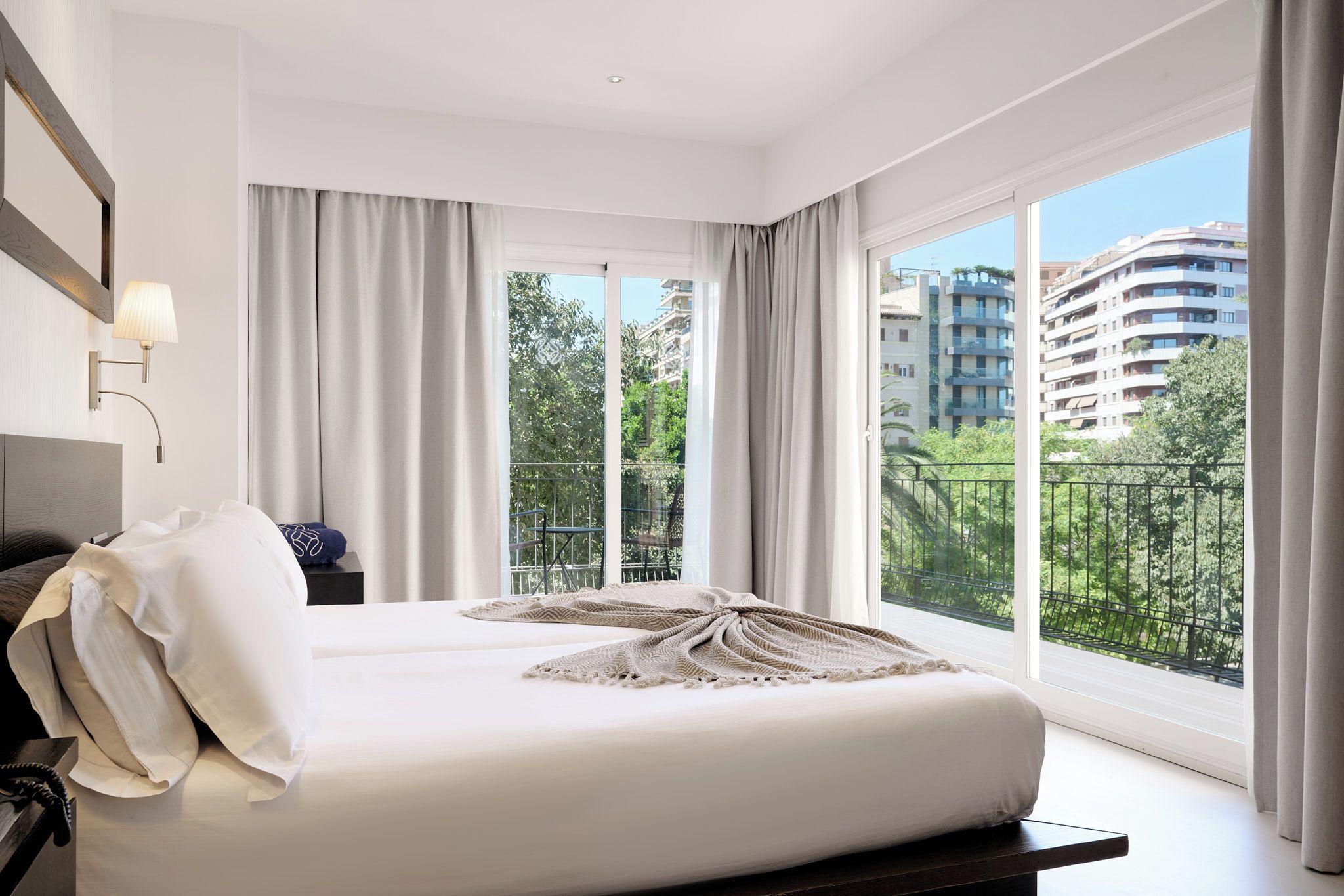 Un dormitorio de hotel moderno con una cama grande, una mesita de noche, una lámpara y vistas de la ciudad a través de amplios ventanales.
