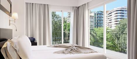 Un dormitorio de hotel moderno con una cama grande, una mesita de noche, una lámpara y vistas de la ciudad a través de amplios ventanales.