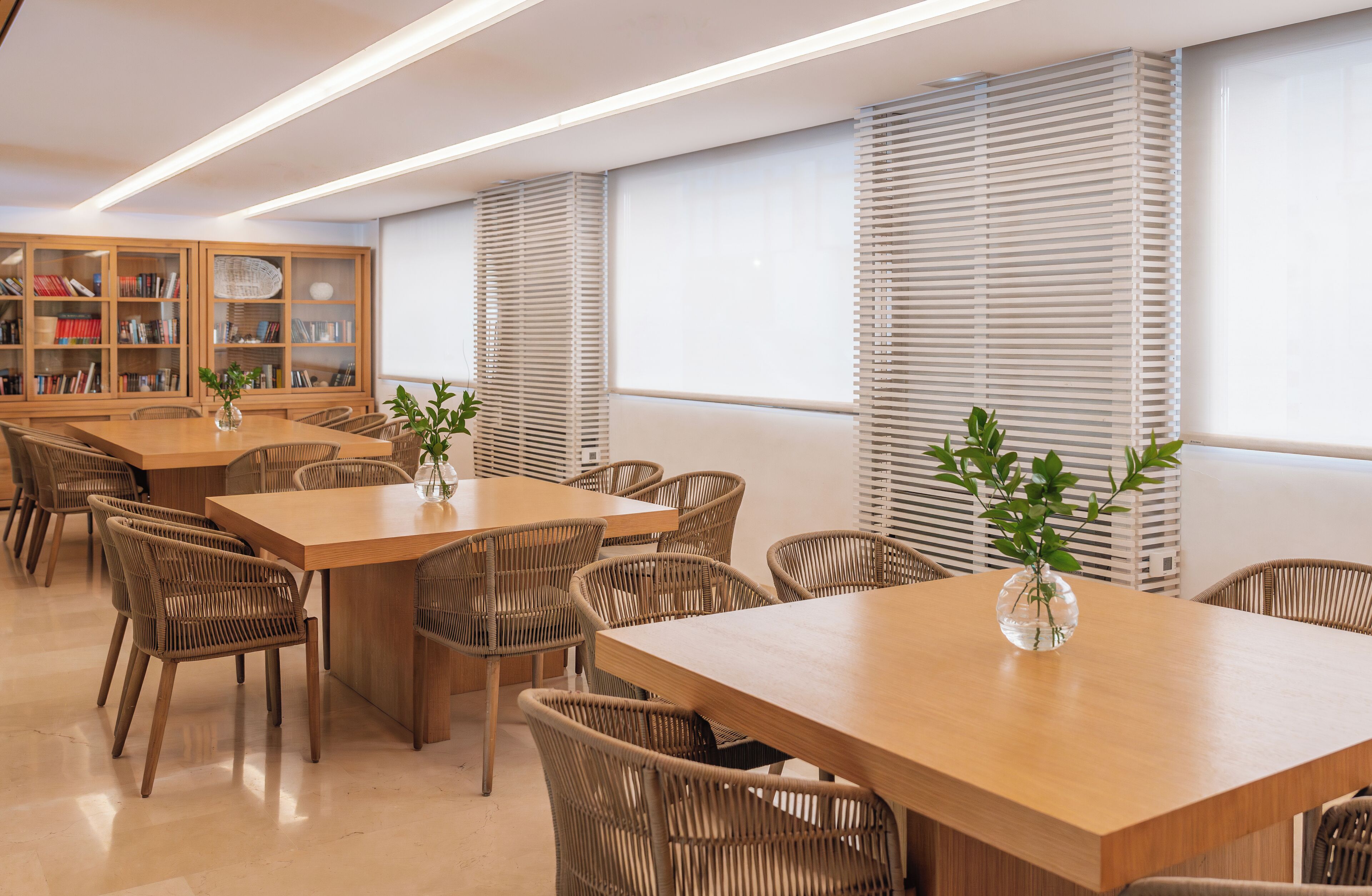 Eine moderne Bibliothek mit Holztischen und Korbstühlen, großen Fenstern mit Jalousien und Bücherregalen voller Bücher.