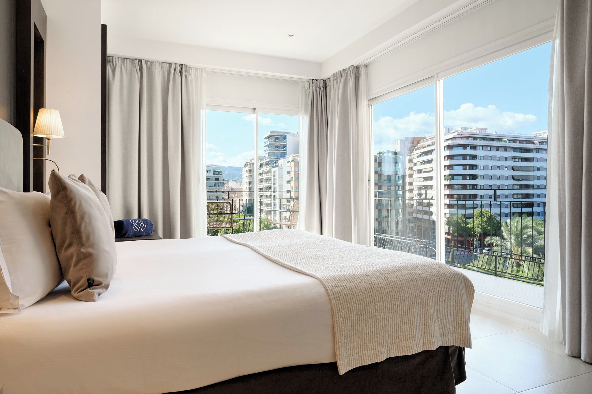 Een hotelkamer met een groot bed, een nachtkastje, een lamp en een uitzicht op de stad door grote ramen.