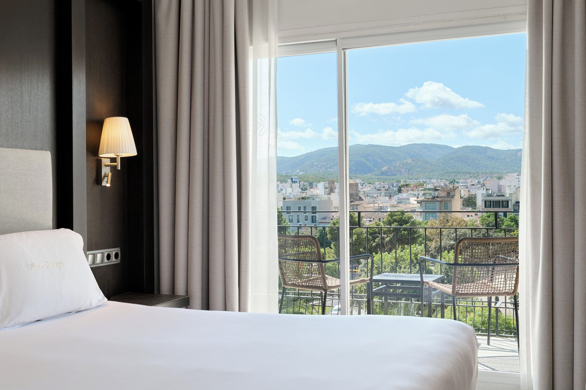 Ein Hotelzimmer mit Bett, Wandlampe und Balkon mit Blick auf Stadt und Berge.