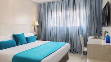 A hotel room with a large bed, a desk with a laptop, a TV, and a window with striped curtains.