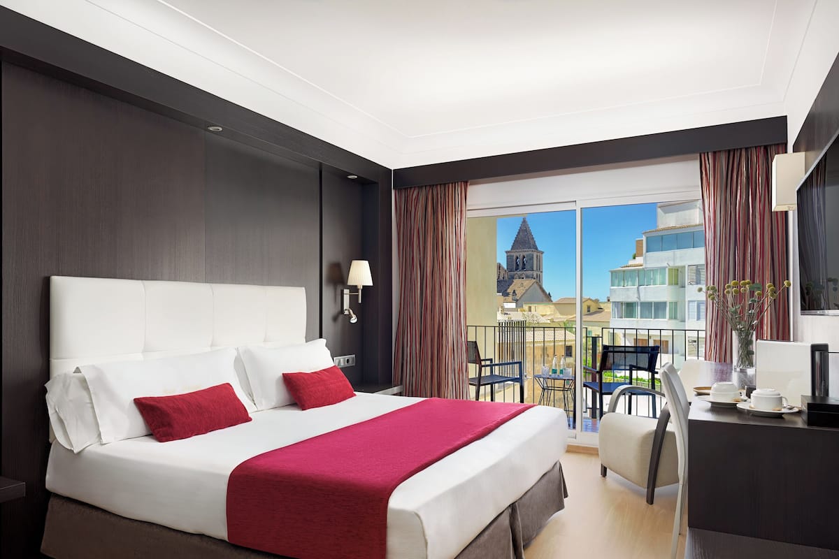 Een hotelkamer met een groot bed, een bureau en een balkon met uitzicht op gebouwen en een toren.