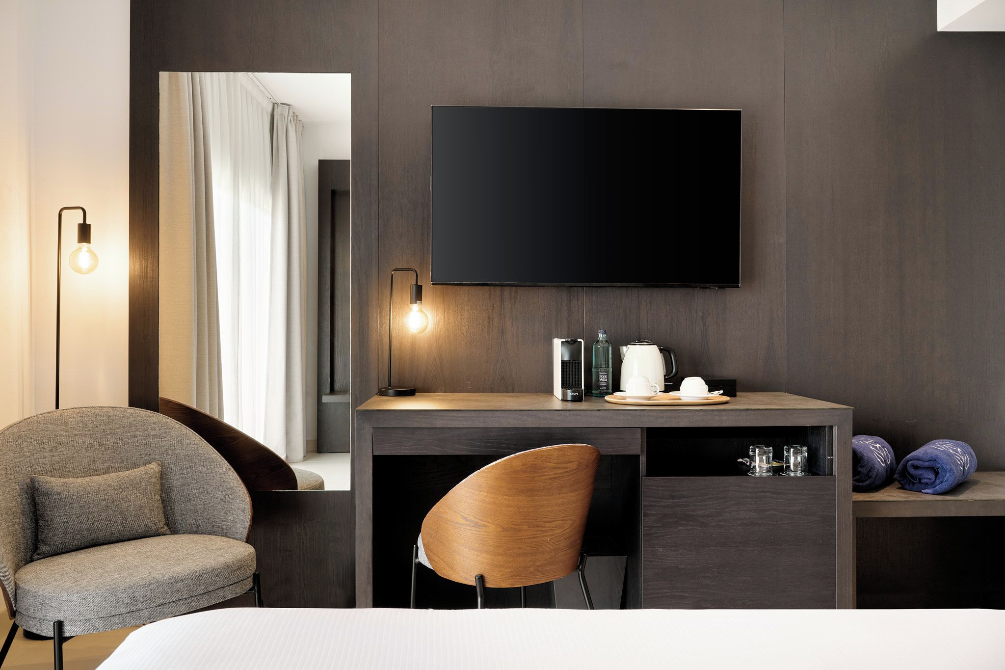 Habitación de hotel con cama, escritorio con televisor de pantalla plana, una silla, mesita de noche con hervidor, tazas y vasos.